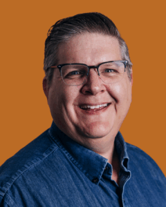 A photo of Austin Campbell wearing a denim shirt with a light brown background.